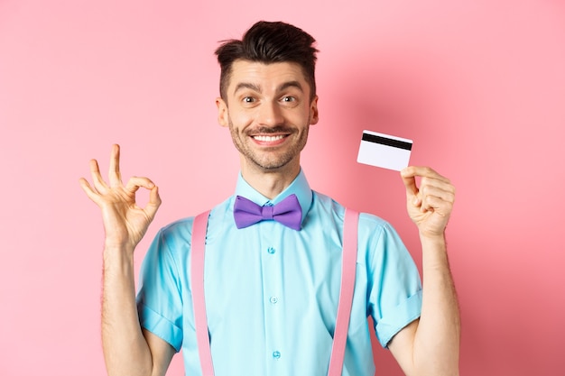 Concepto de compras. Comprador masculino guapo sonriente que muestra el signo de Ok y tarjeta de crédito plástica, comprando algo, de pie satisfecho sobre fondo rosa.