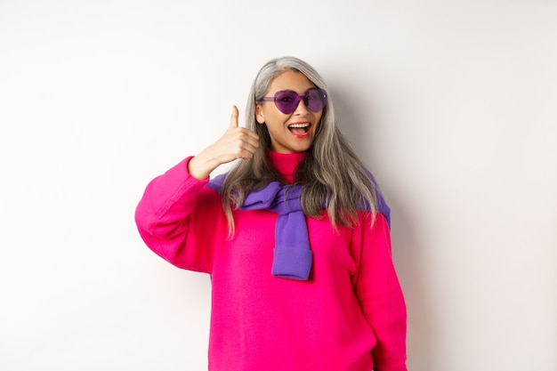 Concepto de compras. Elegante mujer mayor asiática en gafas de sol y ropa de moda, mostrando el pulgar hacia arriba en señal de aprobación, recomendando la tienda, de pie sobre fondo blanco.