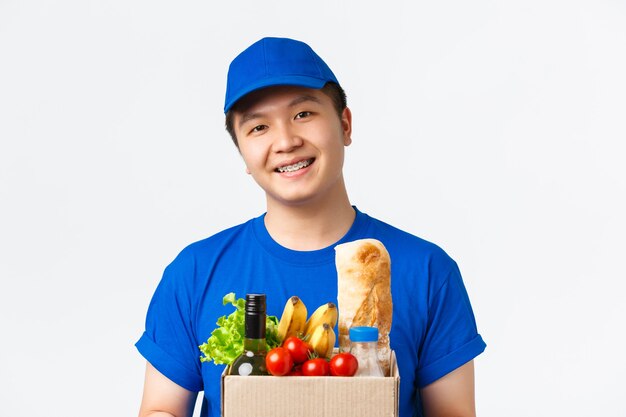 Concepto de compras, entrega de alimentos y envío en línea. Primer plano de mensajero masculino asiático agradable sonriente, vestido con uniforme azul, caja de entrega con pedido de comestibles al cliente, fondo blanco de pie