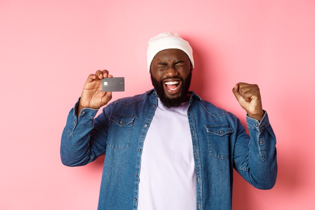 Concepto de compras. Feliz hombre negro regocijándose, gritando de alegría y mostrando la tarjeta de crédito, de pie sobre fondo rosa.