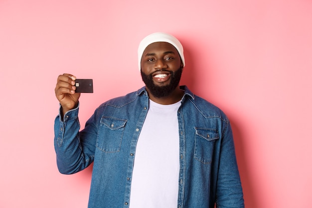Concepto de compras. Hombre barbudo joven satisfecho con gorro mostrando tarjeta de crédito, sonriendo complacido, haciendo compras, de pie sobre fondo rosa.