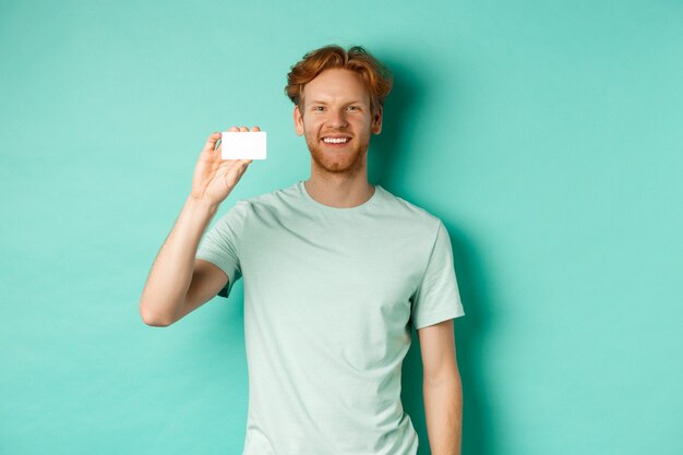 Concepto de compras. Hombre guapo pelirrojo en camiseta mostrando tarjeta de crédito de plástico y sonriendo, de pie sobre fondo turquesa