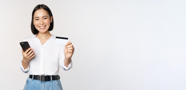 Concepto de compras en línea Una chica asiática moderna sonriente muestra que su tarjeta de crédito tiene un pedido de teléfono móvil con un teléfono inteligente de pie sobre fondo blanco