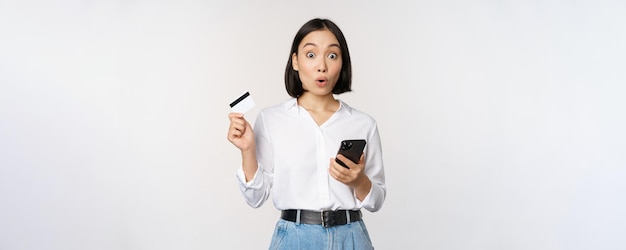 Concepto de compras en línea Imagen de una chica asiática sorprendida sosteniendo una tarjeta de crédito y un teléfono inteligente mirando asombrado con incredulidad ante el fondo blanco de la cámara