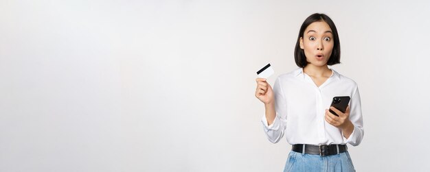 Concepto de compras en línea Imagen de una chica asiática sorprendida sosteniendo una tarjeta de crédito y un teléfono inteligente mirando asombrado con incredulidad ante el fondo blanco de la cámara