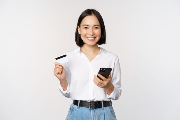 Concepto de compras en línea Imagen de una joven mujer asiática moderna con tarjeta de crédito y compras de teléfonos inteligentes con una aplicación de teléfono inteligente que paga sin contacto de pie sobre fondo blanco