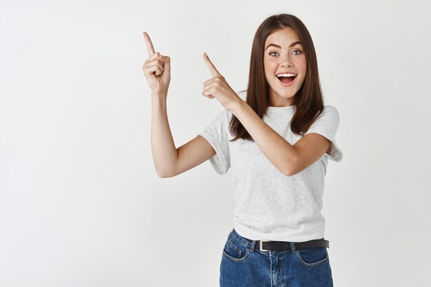 Concepto de compras Mujer joven feliz mostrando el logotipo apuntando a la esquina superior izquierda y sonriendo asombrado de pie sobre fondo blanco.