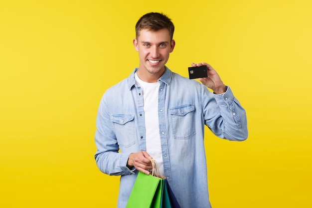 Concepto de compras, ocio y descuentos. Sonriente joven guapo comprando ropa nueva, sosteniendo bolsas y mostrando la tarjeta de crédito con expresión satisfecha, pagando con dinero ahorrado, fondo amarillo.