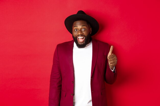 Concepto de compras y personas de Navidad. Guapo hombre negro barbudo en chaqueta de fiesta mostrando el pulgar hacia arriba, mirando sorprendido y satisfecho, fondo rojo.