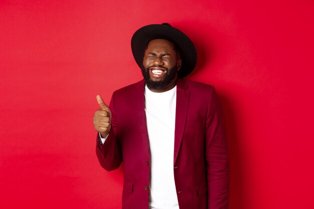 Concepto de compras y personas de Navidad. Guapo hombre negro barbudo en chaqueta de fiesta mostrando el pulgar hacia arriba, riendo y divirtiéndose, de pie sobre fondo rojo.