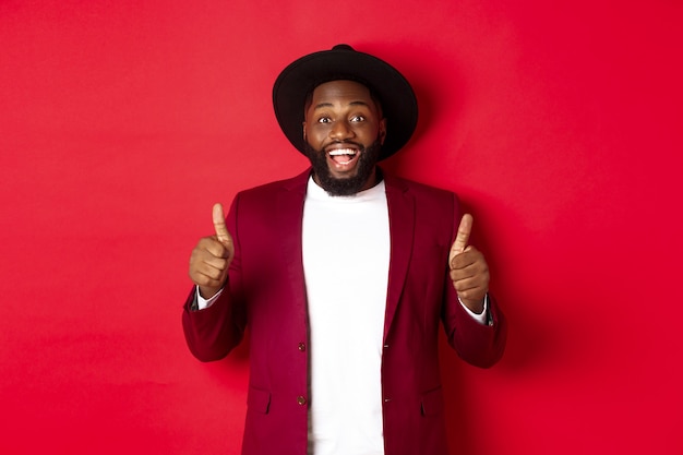 Concepto de compras y personas de Navidad. Hombre negro guapo sonriendo satisfecho, mostrando el pulgar hacia arriba, me gusta y está de acuerdo, aprueba algo, de pie contra el fondo rojo.