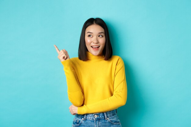 Concepto de compras. Retrato de una atractiva chica coreana en suéter amarillo, mostrando la oferta de promoción en el espacio de la copia, apuntando y mirando hacia la izquierda con una sonrisa de satisfacción, fondo azul
