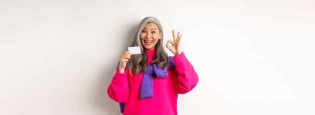 Foto gratuita concepto de compras sonriente mujer asiática de mediana edad con cabello gris mostrando tarjeta de crédito de plástico y ok si