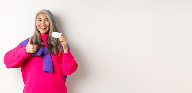 Foto gratuita concepto de compras sonriente mujer asiática de mediana edad con cabello gris mostrando tarjeta de crédito de plástico y pulgar