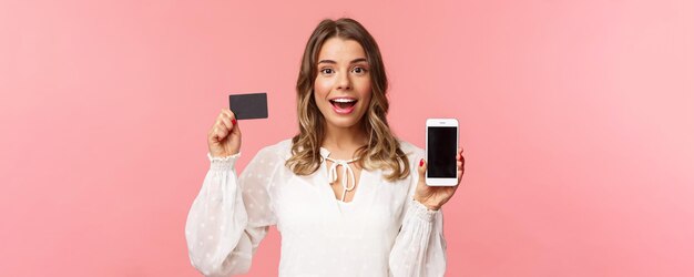 Concepto de compras y tecnología de finanzas Retrato de primer plano de una chica atractiva rubia emocionada con vestido blanco que muestra la tarjeta de crédito y el teléfono móvil anuncian la aplicación de teléfono inteligente de la tienda en línea
