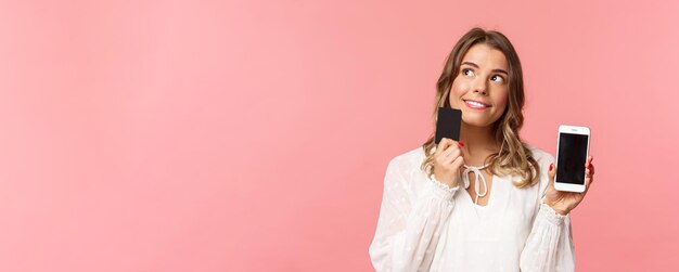 Concepto de compras y tecnología de finanzas Retrato de primer plano de una chica rubia atractiva y soñadora pensativa con vestido blanco pensando qué comprar pedir algo en la tienda en línea sostener el teléfono inteligente y la tarjeta de crédito