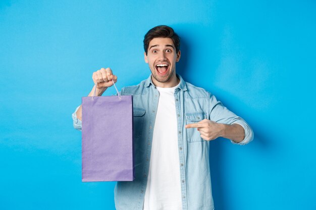 Concepto de compras, vacaciones y estilo de vida. Chico emocionado apuntando con el dedo a la bolsa de papel y mirando asombrado, recomendando la tienda, anunciando descuentos, fondo azul.