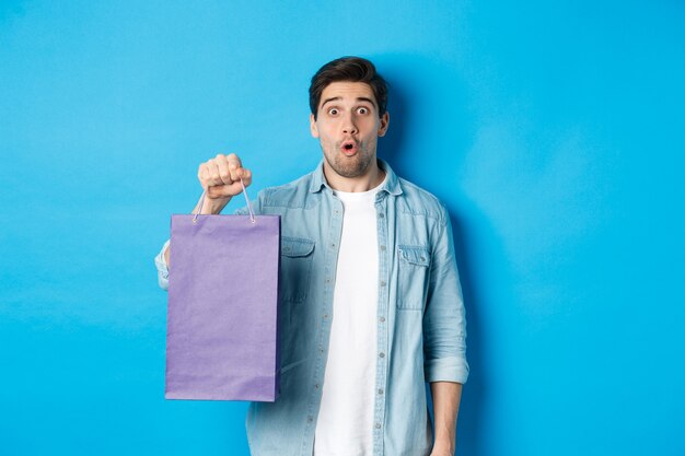 Concepto de compras, vacaciones y estilo de vida. Guapo chico sorprendido sosteniendo una bolsa de papel de la tienda y mirando asombrado, de pie sobre fondo azul.