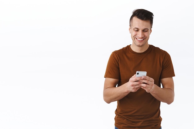 Concepto de comunicación y estilo de vida en línea de tecnología Retrato de un hombre feliz y apuesto que usa un teléfono móvil, mira el teléfono inteligente con una sonrisa radiante, envía mensajes de texto a amigos o compra en Internet