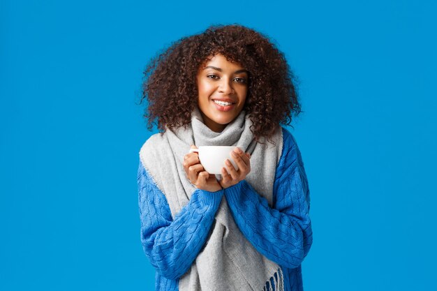 Concepto de confort, ternura y calidez. Encantadora mujer afroamericana encantadora con corte de pelo afro, suéter y bufanda, calentando con una buena taza de té, disfrutando de un café caliente.