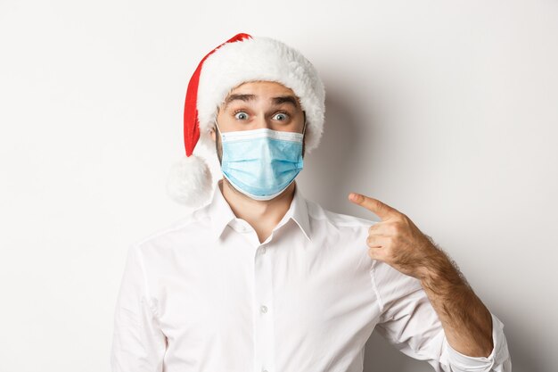 Concepto de coronavirus, cuarentena y vacaciones de invierno. Chico emocionado con sombrero de santa apuntando a su mascarilla, de pie