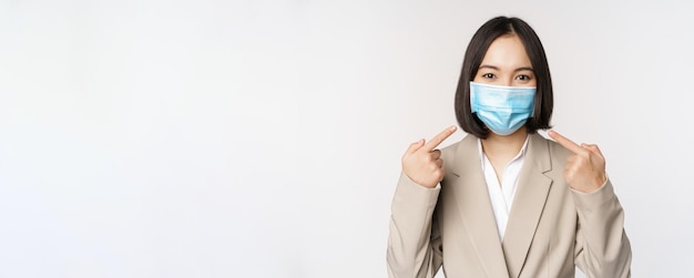 Concepto de coronavirus y gente de negocios Empresaria asiática señalando con el dedo la mascarilla médica en el lugar de trabajo de pie sobre fondo blanco