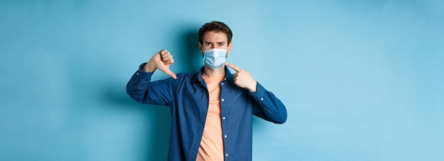 Foto gratuita el concepto de coronavirus y pandemia decepcionó a un joven que señala una máscara médica y muestra el pulgar d