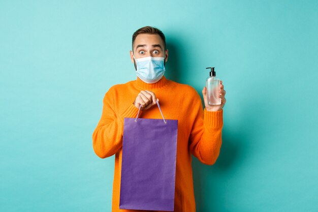 Concepto de coronavirus, pandemia y estilo de vida. Hombre en mascarilla mostrando bolsa de compras y desinfectante de manos