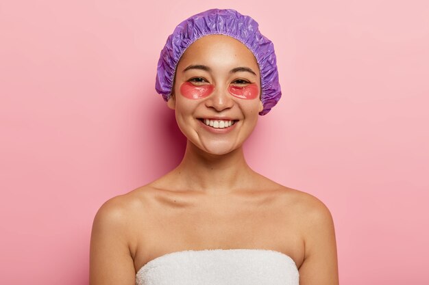 Concepto de cosmetología y cuidado de la piel. Alegre mujer asiática sonríe positivamente, aplica parches de hidrogel debajo de los ojos, usa gorro de ducha, se para sin camisa, envuelta en una toalla blanca, aislada en la pared rosa