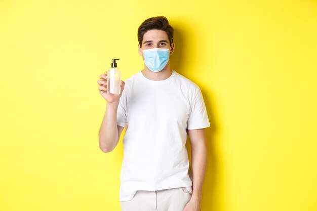 Concepto de covid-19, cuarentena y estilo de vida. Hombre joven en máscara médica mostrando desinfectante de manos, producto de desinfección de manos, de pie sobre fondo amarillo.