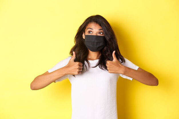 Foto gratuita concepto de covid-19, distanciamiento social y estilo de vida. imagen de una alegre niña afroamericana con mascarilla negra, mostrando el pulgar hacia arriba y mirando feliz en la esquina superior izquierda, fondo amarillo.