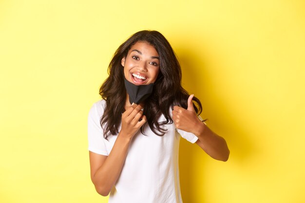 Concepto de covid-19, distanciamiento social y estilo de vida. Retrato de atractiva mujer afroamericana sonriendo, quitándose la mascarilla y mostrando el pulgar hacia arriba, recomiendo algo.