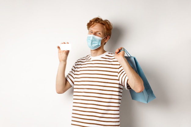 Concepto de covid-19 y estilo de vida. Comprador joven feliz en mascarilla sosteniendo la bolsa de compras y mostrando la tarjeta de crédito de plástico, comprando con descuentos, fondo blanco.