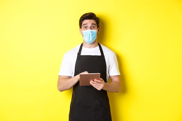 Concepto de covid-19, pequeña empresa y pandemia. Camarero en delantal negro y máscara médica tomando orden, sosteniendo tableta digital, de pie sobre fondo amarillo.