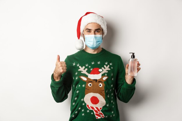 Concepto de covid-19 y vacaciones navideñas. Hombre recomendando desinfectante de manos, mostrando el pulgar hacia arriba y antiséptico, con máscara médica con gorro de Papá Noel, fondo blanco.