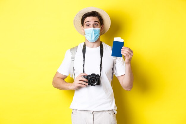 Concepto de covid-19, viaje y cuarentena. Turista hombre feliz con cámara, mostrando pasaporte y boletos para vacaciones, yendo de viaje durante la pandemia, fondo amarillo