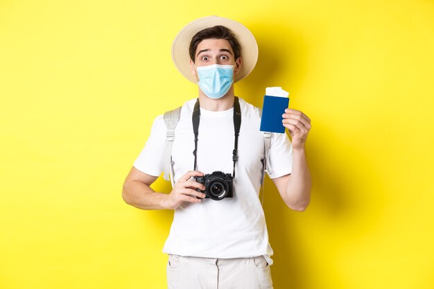 Concepto de covid-19, viaje y cuarentena. Turista hombre feliz con cámara, mostrando pasaporte y boletos para vacaciones, yendo de viaje durante la pandemia, fondo amarillo.