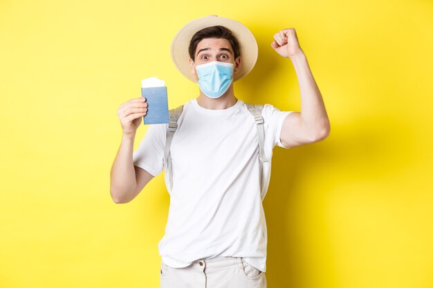 Concepto de covid-19, viaje y cuarentena. Turista de hombre feliz con máscara médica celebrando, mostrando el pasaporte con boletos para vacaciones y regocijo, viaje durante el coronavirus.