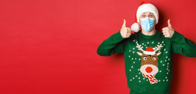 Concepto de covid de año nuevo y distanciamiento social hombre guapo alegre con sombrero de santa y máscara médica ...