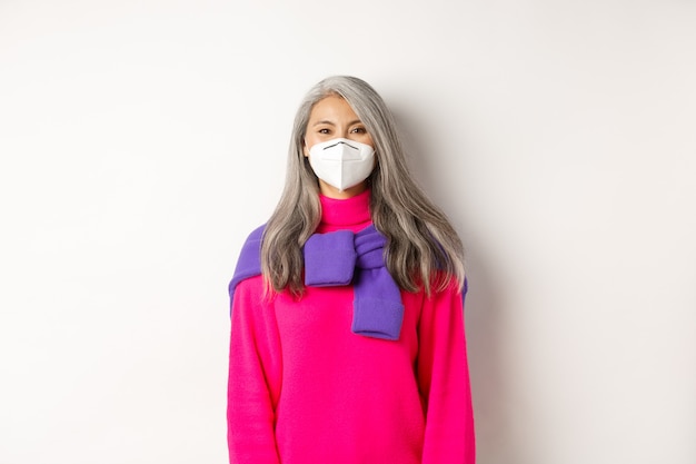 Concepto de covid, pandemia y distanciamiento social. Alegre modelo de mujer senior asiática en mascarilla sonriendo a la cámara, medidas preventivas del coronavirus, fondo blanco.