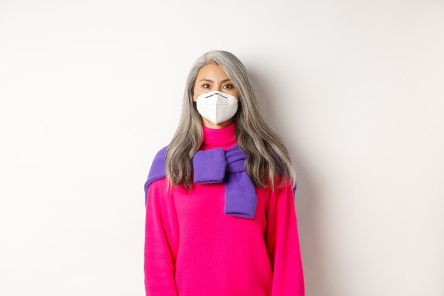 Concepto de covid, pandemia y distanciamiento social. Elegante mujer asiática senior con respirador y mirando a la cámara seria, de pie sobre fondo blanco.