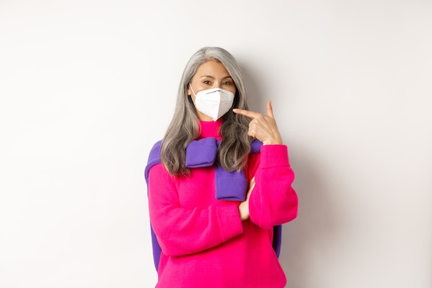 Concepto de covid, pandemia y distanciamiento social. Mujer mayor asiática de moda con respirador, apuntando a la mascarilla y sonriendo, de pie sobre fondo blanco.