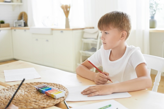 Concepto de creatividad, ocio, afición, arte e imaginación. Imagen de colegial caucásico pensativo en camiseta blanca sentado en el escritorio en el interior, con mirada pensativa, pensando qué dibujar con lápiz