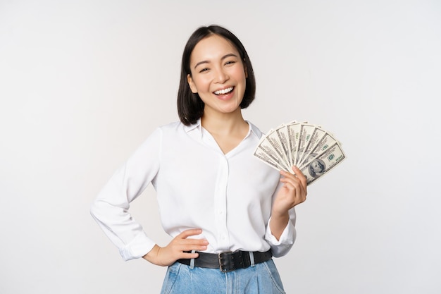 Concepto de crédito y préstamo Mujer asiática joven sonriente sosteniendo dólares en efectivo y mirando feliz el fondo blanco de la cámara