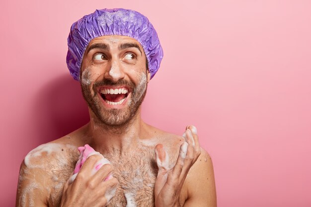 Concepto de cuidado corporal. Chico feliz se ducha, se relaja después del trabajo estresado, tiene la piel húmeda con espuma, se frota el pecho con una esponja, mira con alegría a un lado, aislado en la pared rosa