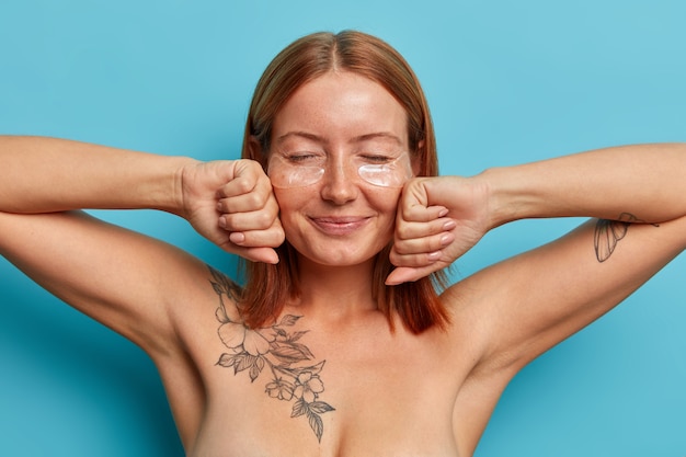 Concepto de cuidado personal y cosmetología. mujer pecosa complacida con cabello pelirrojo natural, mantiene las manos en el puño cerca de la cara, se para desnuda contra la pared azul, usa parches debajo de los ojos para levantar