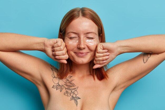 Concepto de cuidado personal y cosmetología. Mujer pecosa complacida con cabello pelirrojo natural, mantiene las manos en el puño cerca de la cara, se para desnuda contra la pared azul, usa parches debajo de los ojos para levantar