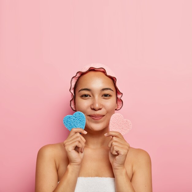 Concepto de cuidado de la piel. Hermosa chica coreana satisfecha sostiene esponjas cosméticas rosadas y azules en forma de corazón, limpia la cara, elimina los poros, quiere lucir renovada, usa una toalla blanca