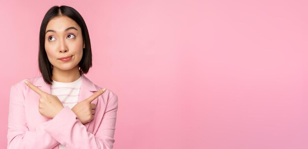 Concepto de decisión Joven empresaria asiática trabajadora corporativa señalando con el dedo hacia los lados en dos direcciones que muestran variantes que parecen indecisas al elegir un fondo rosa
