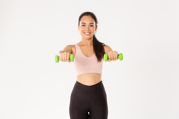 Concepto de deporte, bienestar y estilo de vida activo. Alegre sonriente chica asiática fitness, deportista levantando mancuernas, entrenamiento en los músculos, ganando bíceps con ejercicios en casa, fondo blanco.